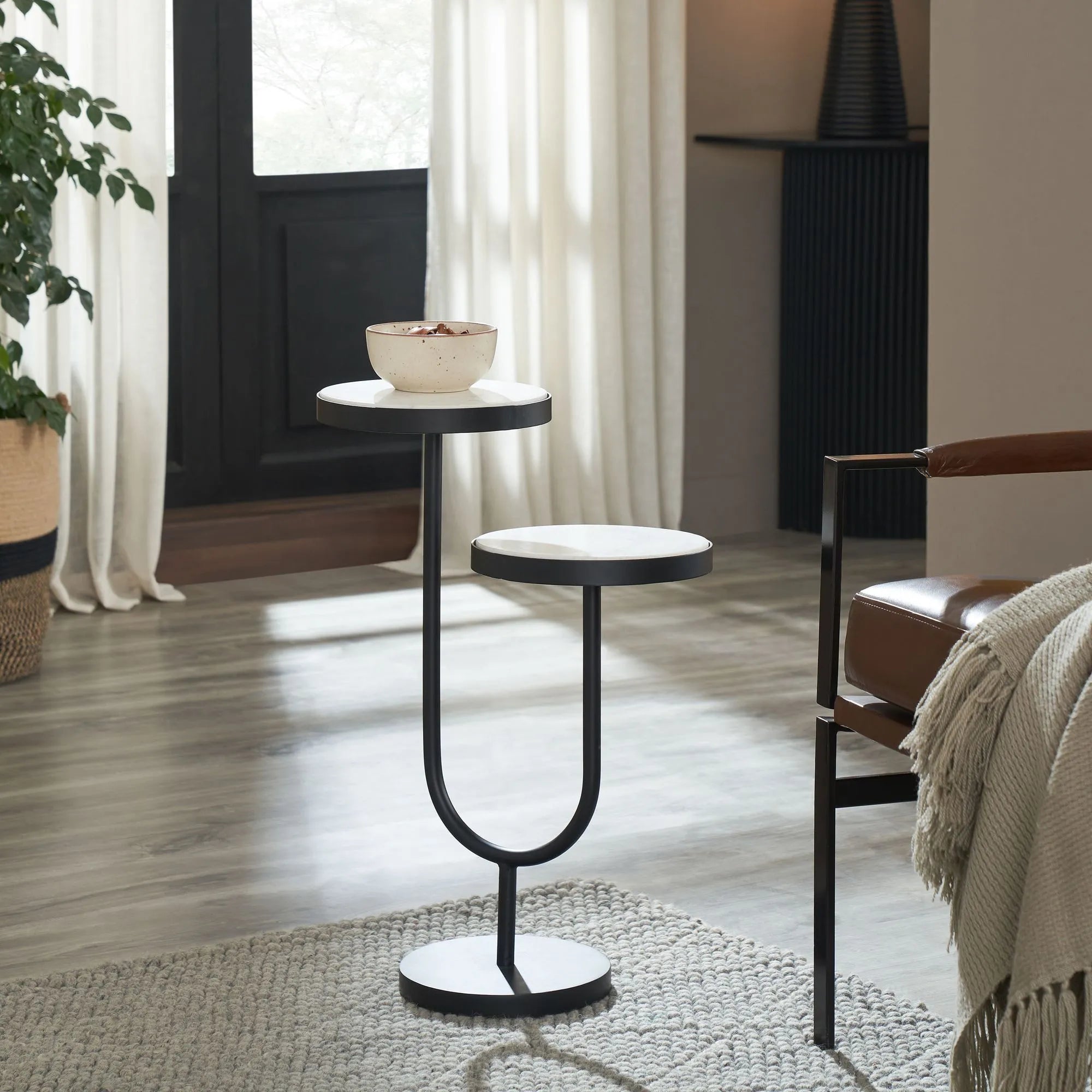 The Ambra is a modern black iron table with two round white marble tops at different heights, set on a wooden floor by a brown leather chair, complemented by a knit blanket and a potted plant by the window.