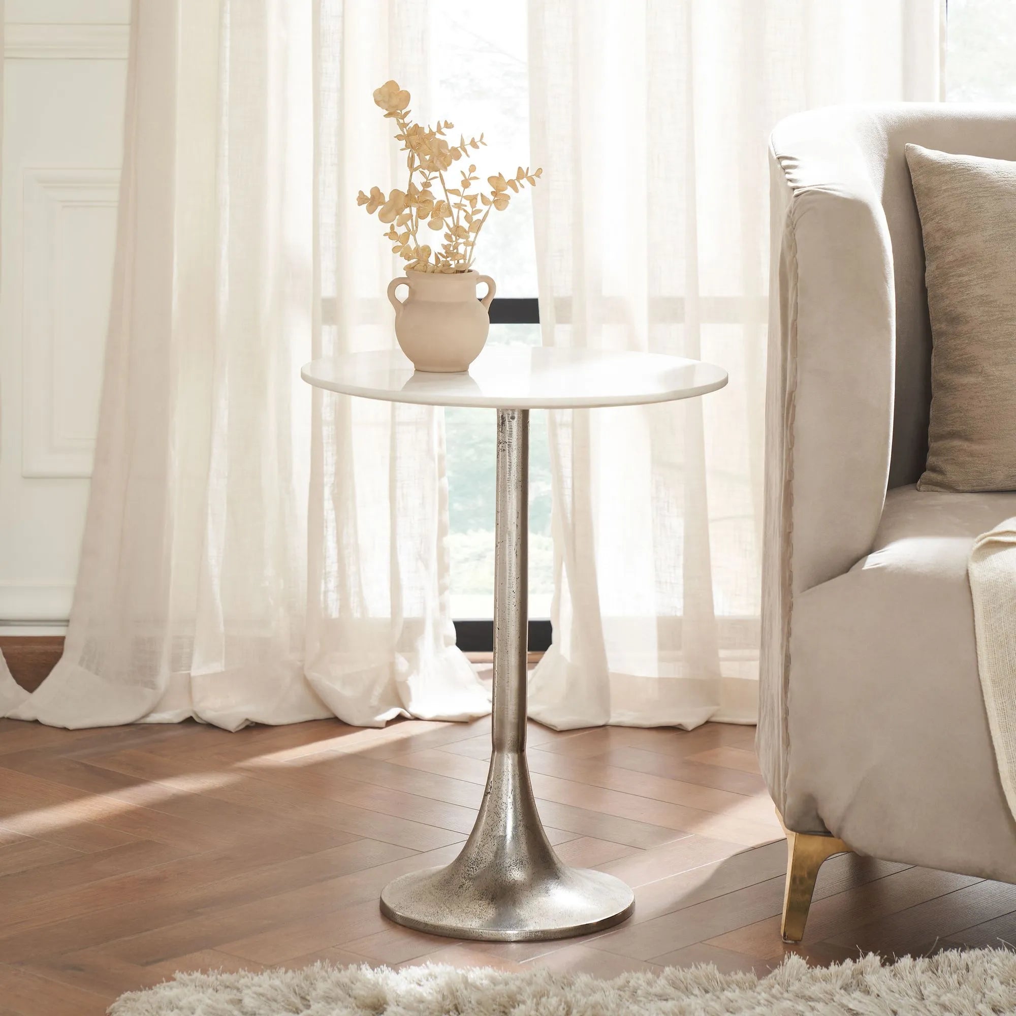 The Eloa side table, featuring a marble top and antique nickel tulip base, holds a beige vase with dried flowers beside a light-colored sofa in a sunlit room with sheer curtains and wooden floors.