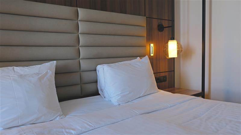 A bed with white linens and two pillows rests against a Custom Upholstered Headboard. A bedside table with a modern lamp on the right completes the clean, inviting look.
