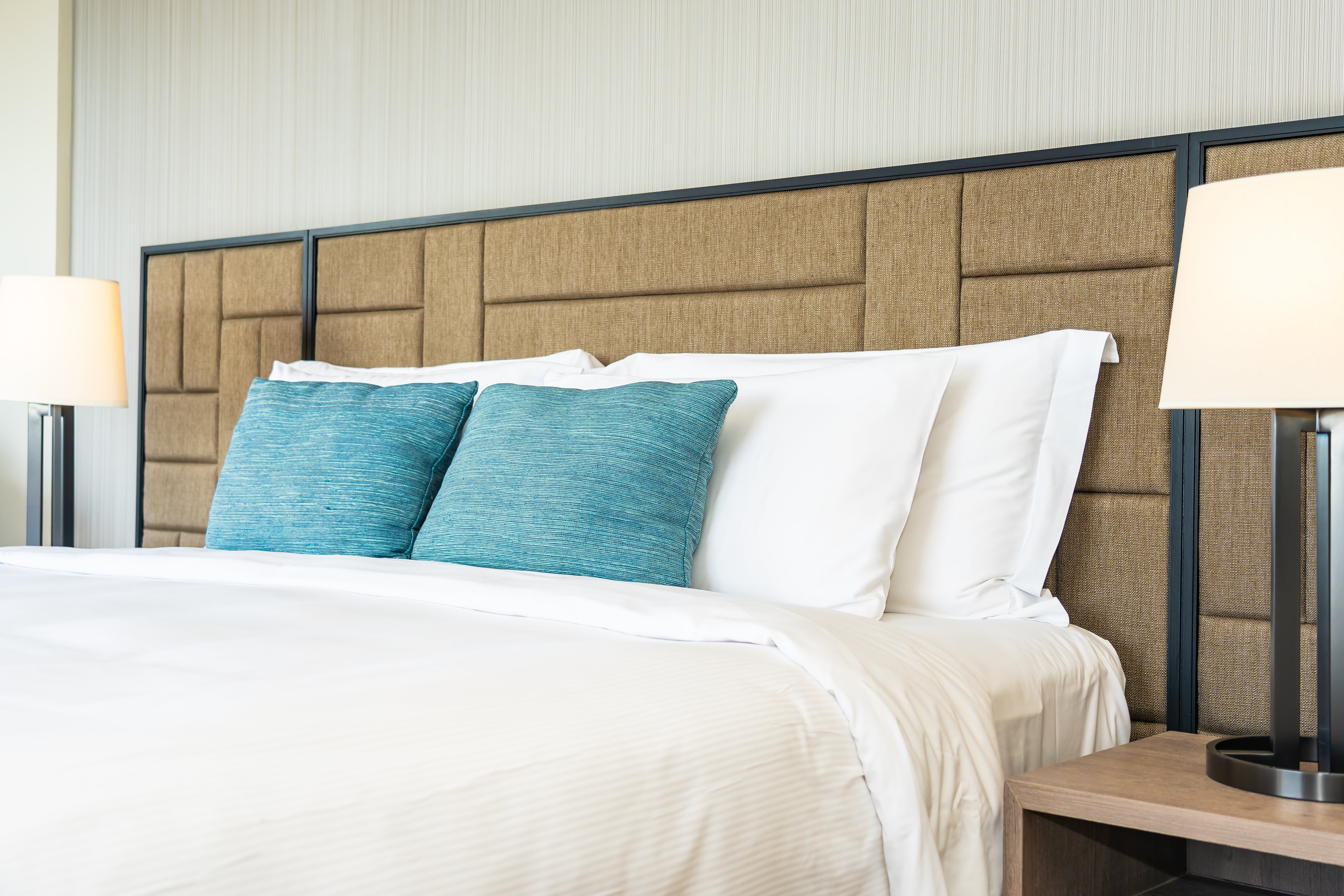 A bed with white sheets, teal accent pillows, and a large Custom Upholstered Headboards piece. Modern table lamps are placed on wooden nightstands beside the bed.