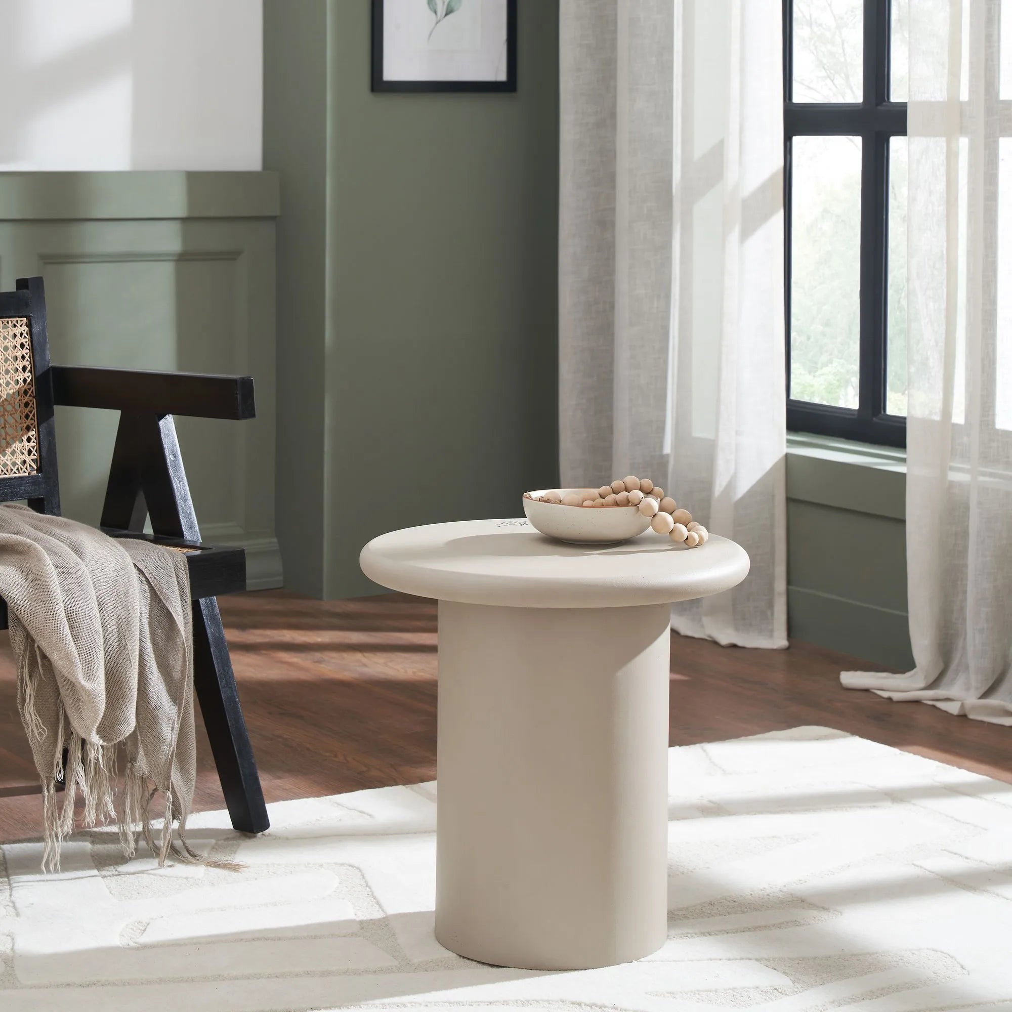 A minimalist living room features the Riya round beige accent table with a bowl of wooden beads, next to a black chair draped in a beige throw, by a window with sheer curtains and green walls.