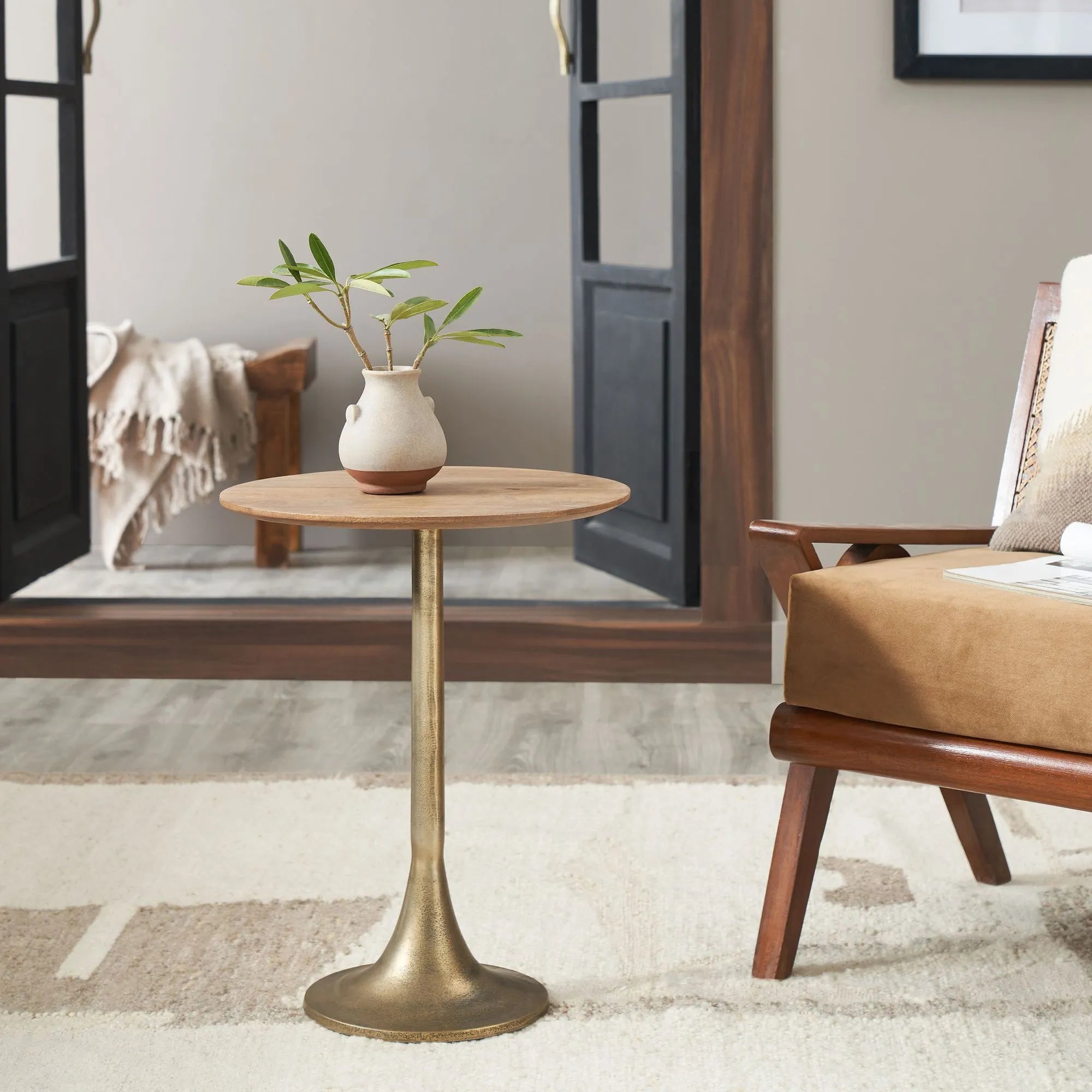 The Sparta, a small round mango wood side table with an antique brass tulip base, displays a ceramic vase with green branches. It sits by a tan armchair on a light rug, with a mirror and blanket in the background.