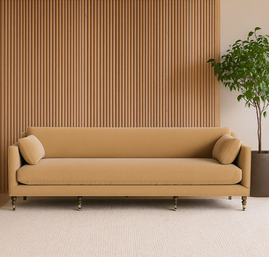 A modern living room showcases the tan Strathmore Sofa with two round pillows, placed before a vertical wood slat wall. A green potted plant on beige carpet to the right adds a welcoming, hospitality-inspired touch.